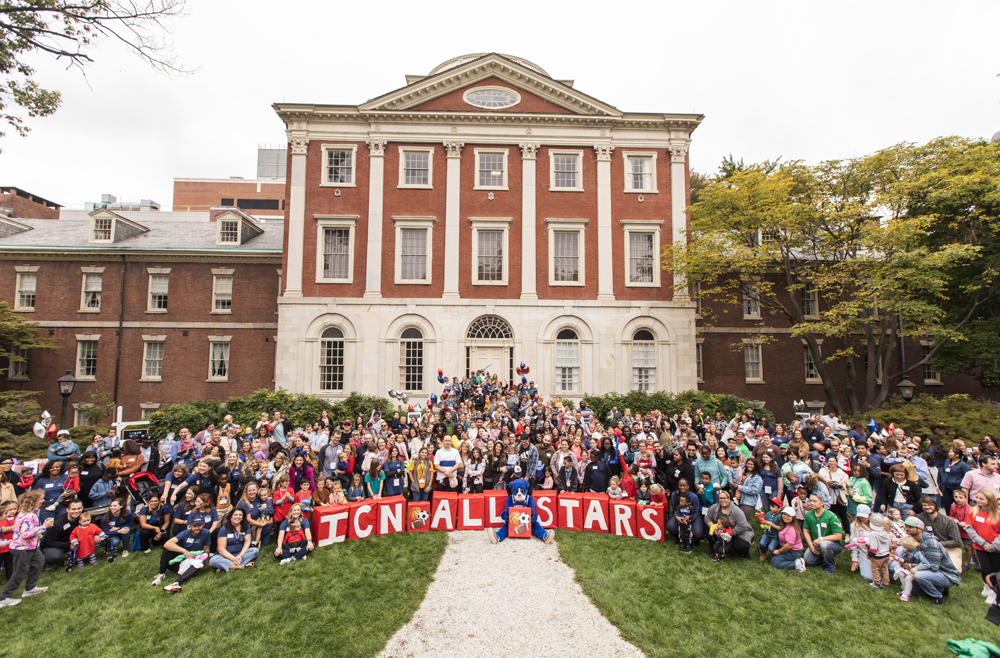 A large group of families with children, joined by staff, pose for a photo in Pennsylvania Hospital's Elm Garden, standing behind big red boxes that read, "ICN All Stars."